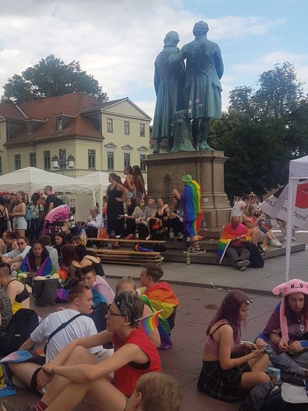 Auf einem Platz sitzen viel junge Menschen, die teilweise eine Regenbogen-Fahne umgehängt haben. In der Mitte sieht man befindet sich die Statue von Goethe und Schiller 